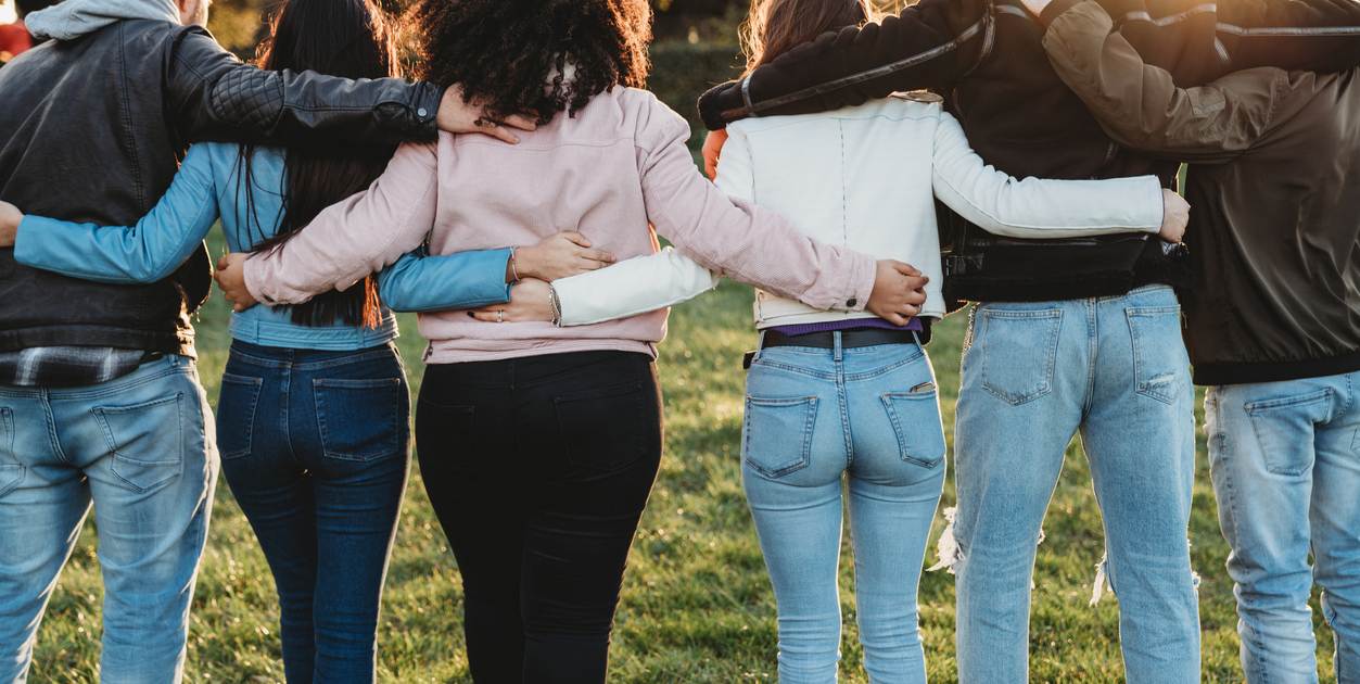 Group of six teenager friends embracing together at the park, rear view
